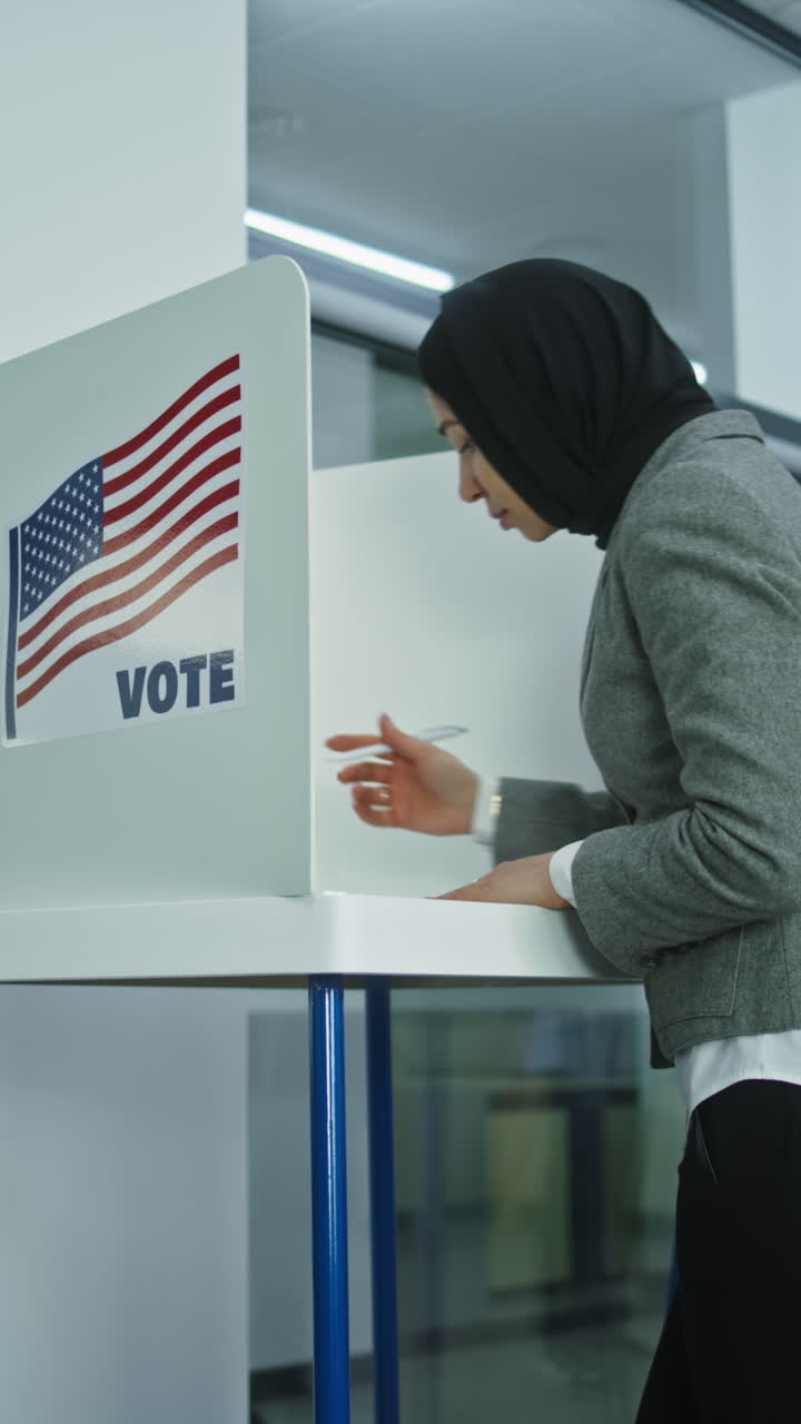 Muslim woman in hijab comes to vote in booth in polling station office. National Election Day in the United States. Political races of US presidential candidates. Concept of civic duty. Dolly shot.