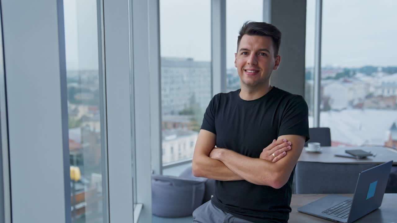 Millennial male wearing casual clothes stands leaning on desk. Young man folding hands on his chest looking at camera. Close up portrait.