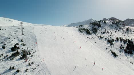 drone rises laterally across a ski slope in andorra as skiers descend, revealing blue sky above.