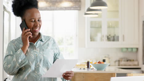 african american senior woman doing paperwork and using smartphone in sunny room, slow motion