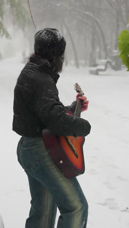 vista lateral de una mujer joven con capucha negra tocando la guitarra acústica en un parque cubierto de nieve rodeado de árboles helados, creando una atmósfera serena de invierno
