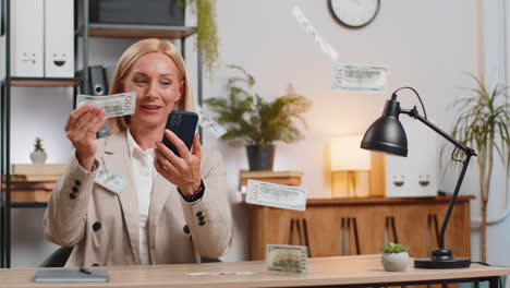 money cash dollar rain falling on mature businesswoman using smartphone celebrating success at desk