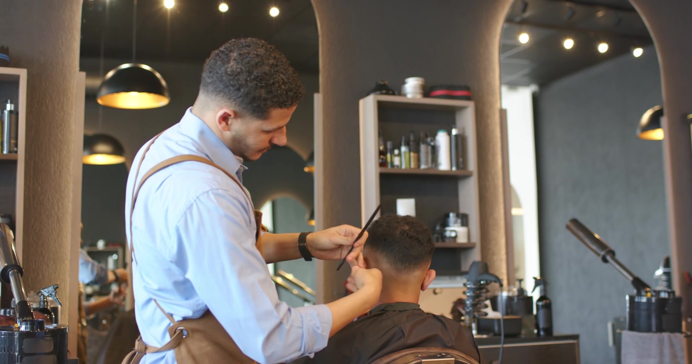 Afroamerikansk barber og kunde sidder og klipper/trimmer hår til en fade på salon med trimmer