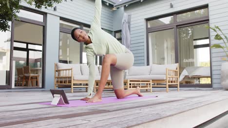 mujer biracial enfocada practicando yoga y estirándose con una tableta en el jardín, cámara lenta