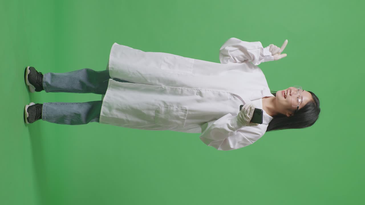 연구실의 녹색 화면 배경에 서있는 동안 스마트폰으로 미소 짓고 을 가리키는 아시아 여성 과학자의 전체 몸