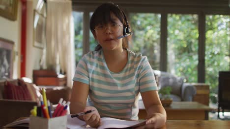 Asian-woman-sitting-at-table-wearing-headphones-and-making-video-call