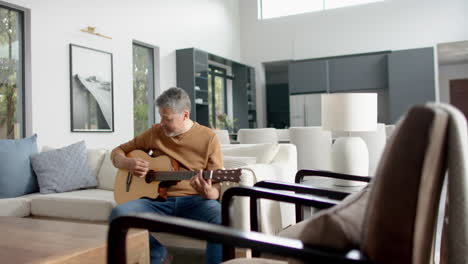 senior biracial man sitting on couch playing guitar at home, copy space, slow motion