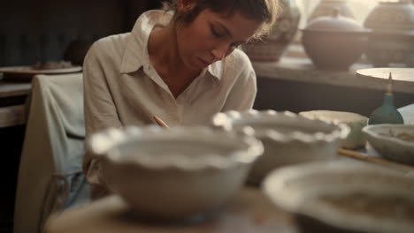 serious woman scraping clay plate in pottery. girl carving on product in studio