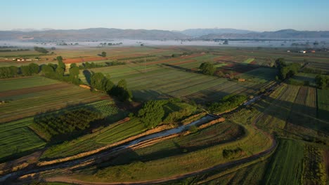 Vista-Aérea-De-Parcelas-Agrícolas-Verdes-Alrededor-De-Una-Granja-Europea-Al-Amanecer-En-Primavera,-Vuelo-Matutino