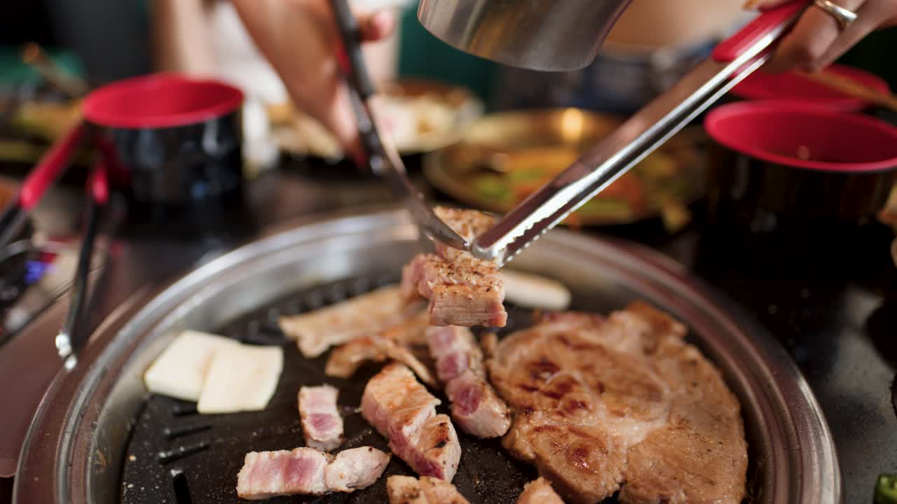 Person cuts pork belly on tabletop grill using tongs and scissors, indoor restaurant, natural lighting