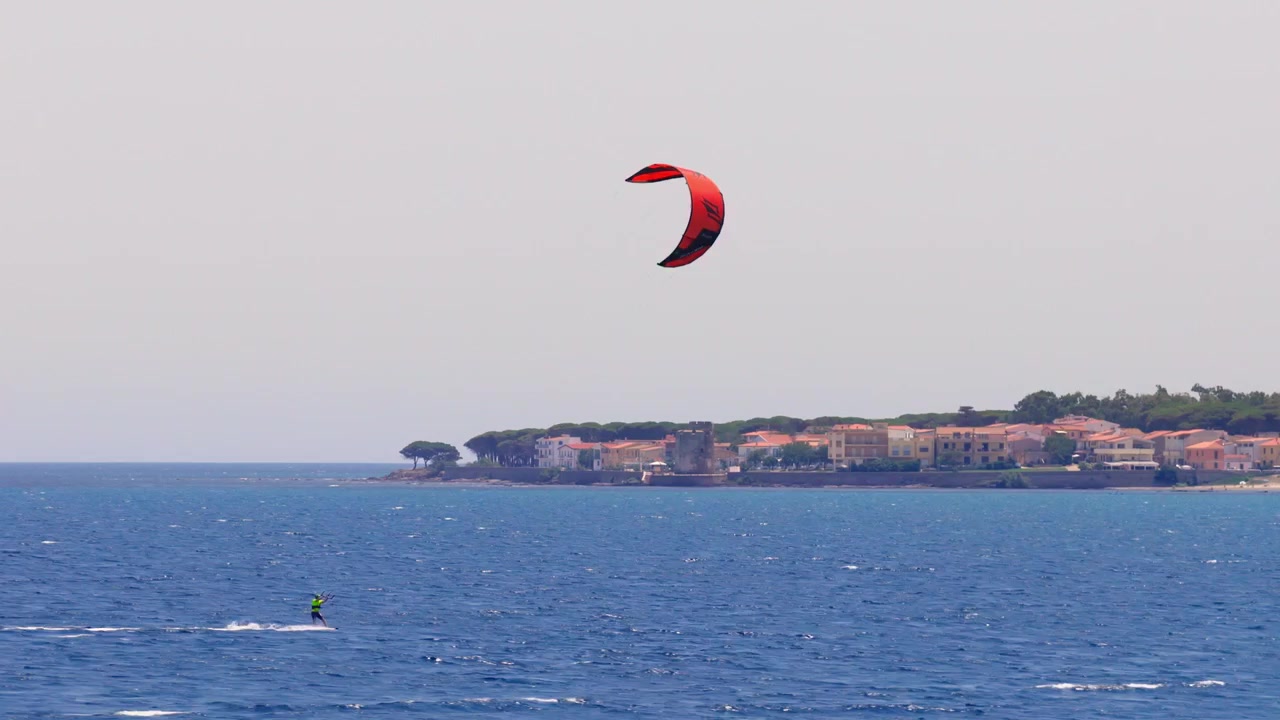 A kitesurfer throws a large fan of spray during a hard carving turn across deep blue water, at La Caletta in Sardinia, Italy.