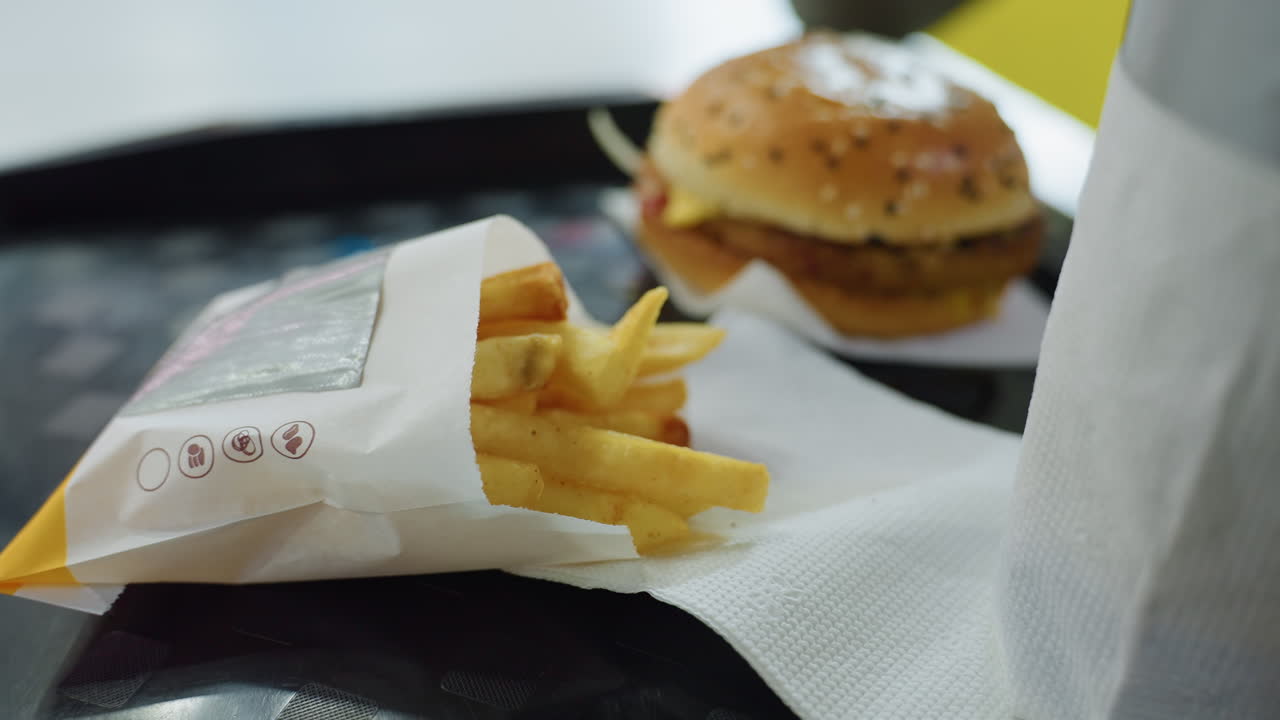 Close up of hand picking french fries from paper bag on tray with burger in background on white napkin during breakfast at indoor fast food restaurant with soft daylight and clean atmosphere
