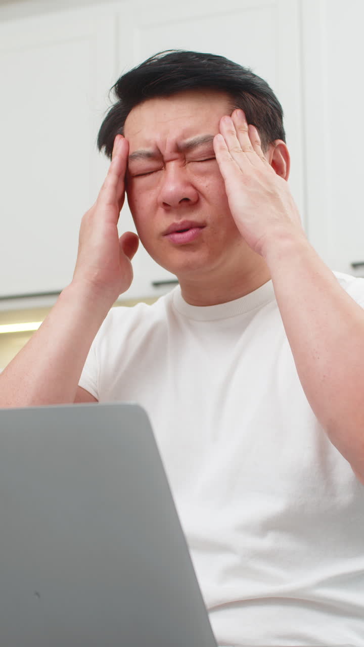 Asian man with laptop feels headache rubs temples closes eyes breathes slowly takes short work break