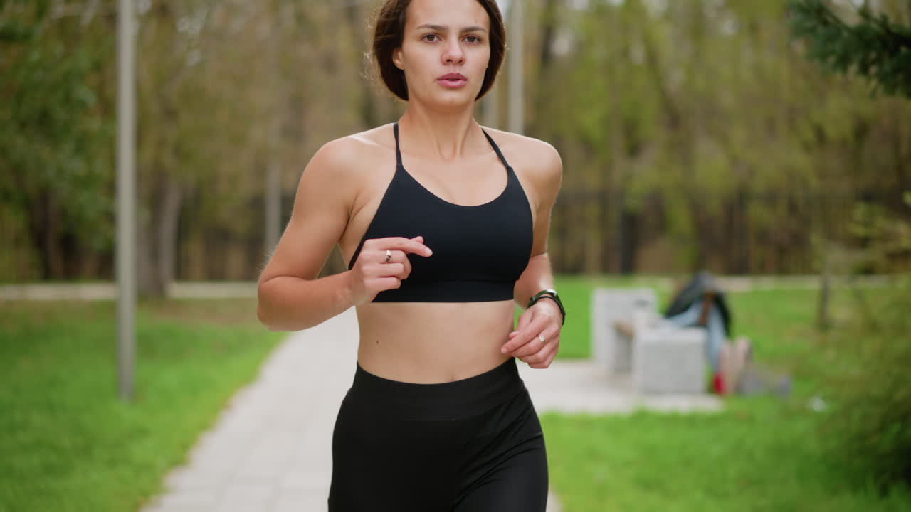 Primo piano di donna atletica in abbigliamento sportivo nero che corre nel parco, con sfondo sfocato che mostra panchina in cemento, pali, alberi e recinzione in ferro, ideale per fitness, jogging e attività all'aperto