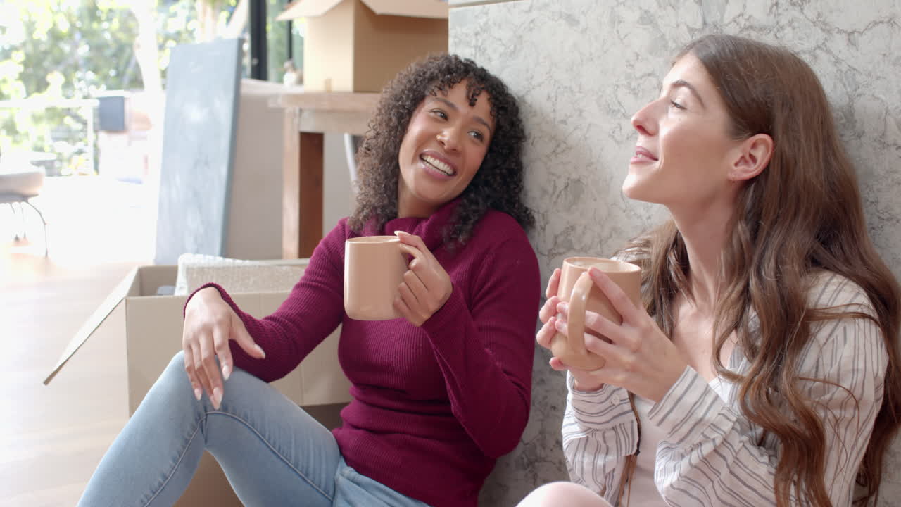 Relaxing with coffee mugs, lesbian couple sitting on floor in new home