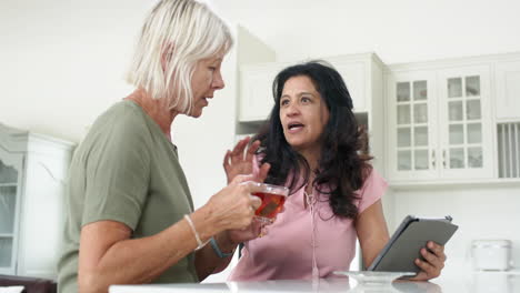 Happy-diverse-senior-women-using-tablet-and-drinking-tea-in-sunny-kitchen,-slow-motion