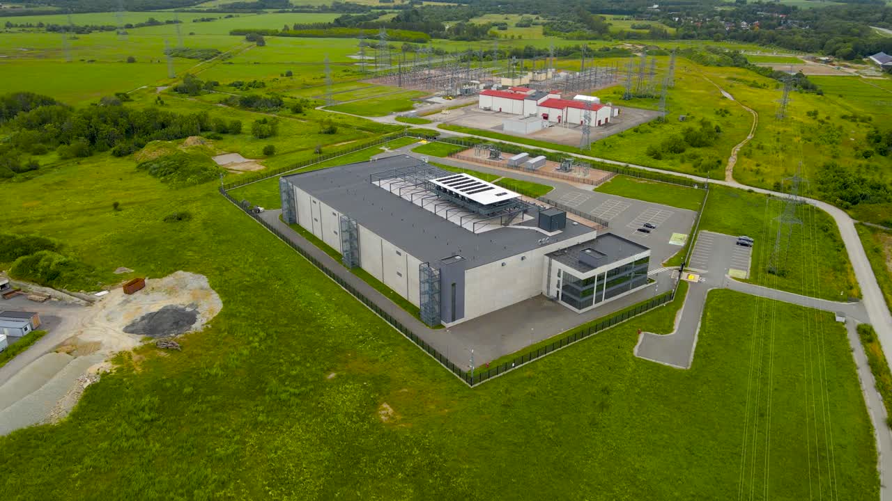 Aerial drone footage orbiting and spinning around a large modern gray or grey colored data centre in green countryside at europe with a power station with electrical lines and wires in the background
