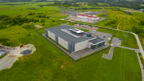 aerial drone footage orbiting and spinning around a large modern gray or grey colored data centre in green countryside at europe with a power station with electrical lines and wires in the background