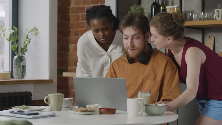 Junge zeigt und erklärt seinen beiden weiblichen Mitbewohnerinnen in der Küche etwas am Laptop