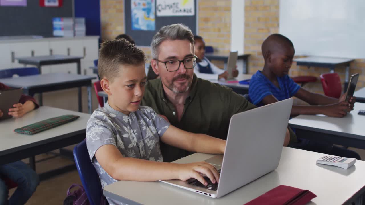 Diverse male teacher helping a schoolboy sitting in classroom using laptop