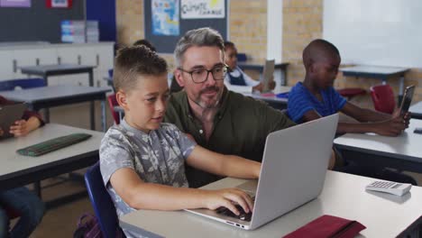 diverse male teacher helping a schoolboy sitting in classroom using laptop