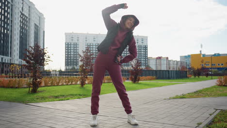 excited female coach in hoodie and black vest stretches side outdoors under sunny sky on urban plaza with tall apartment towers and autumn shrubs during morning workout session by pavement
