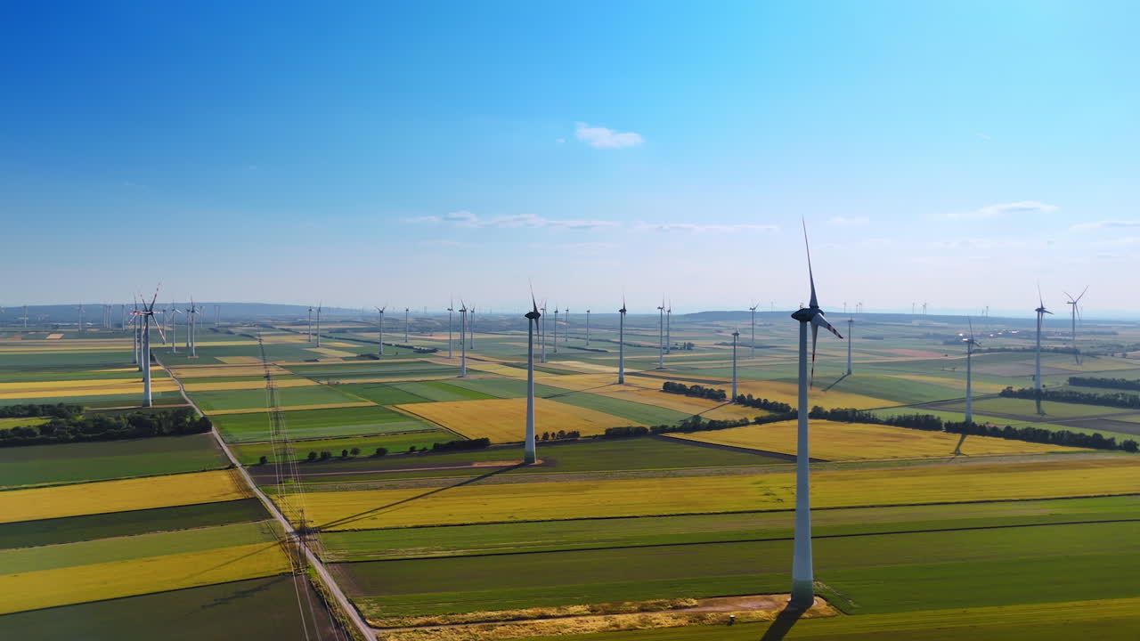 Beautiful patched agricultural plantation with wind farm. Blades of windmills rotate slowly in the field. Aerial perspective.