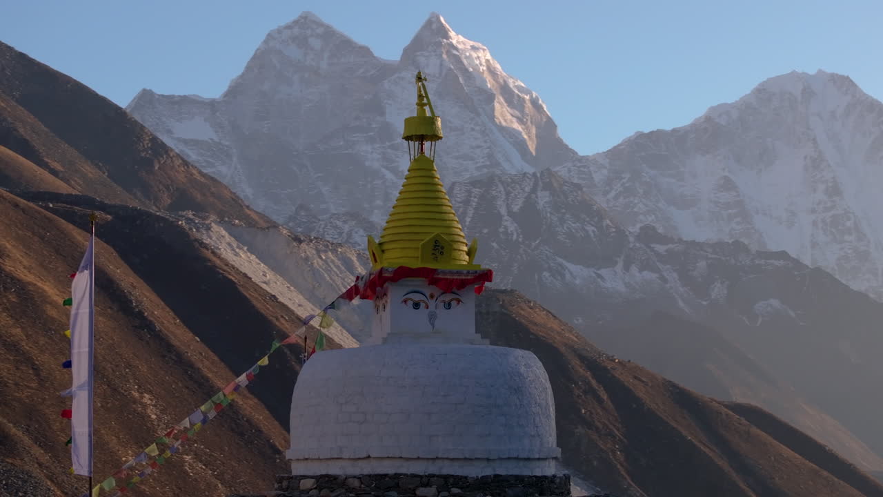 Orbiting aerial drone shot of a Buddhist stupa at Dingboche along the Everest Base Camp trek in Nepal. Snow-capped Himalayan mountains glow during golden hour as prayer flags flutter