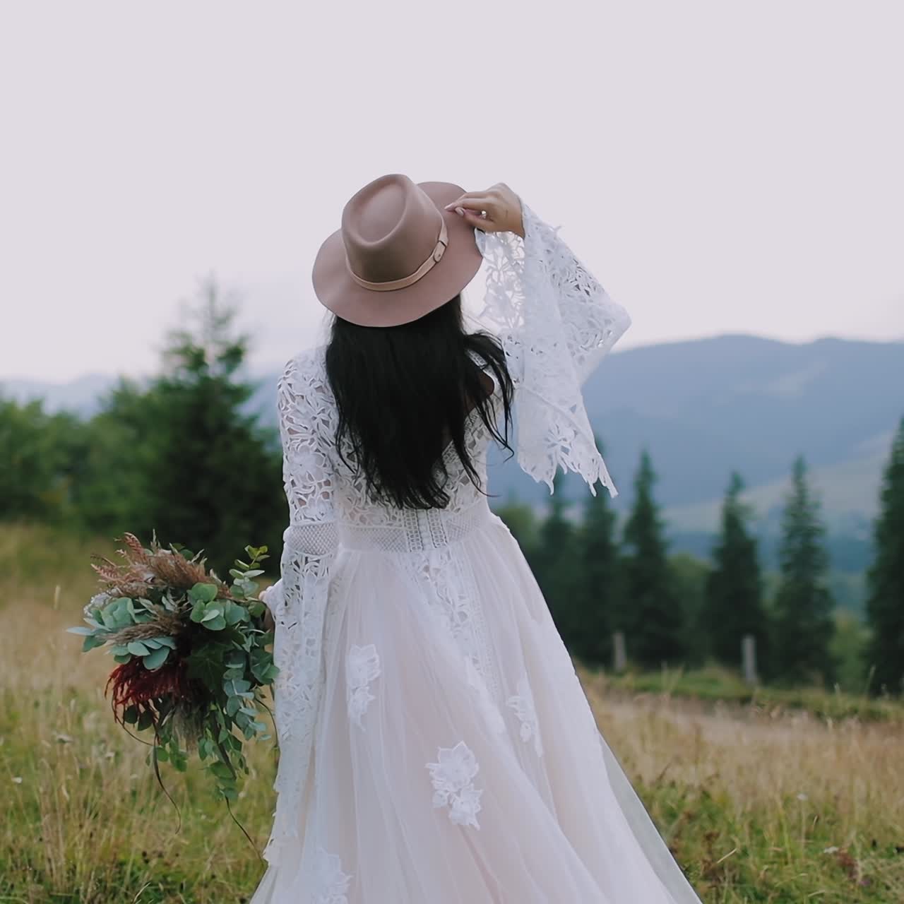 Rear view of a bride in the mountains. Girl in wedding dress and pink hat holding bouquet of flowers and looking on the beautiful mountain landscape.