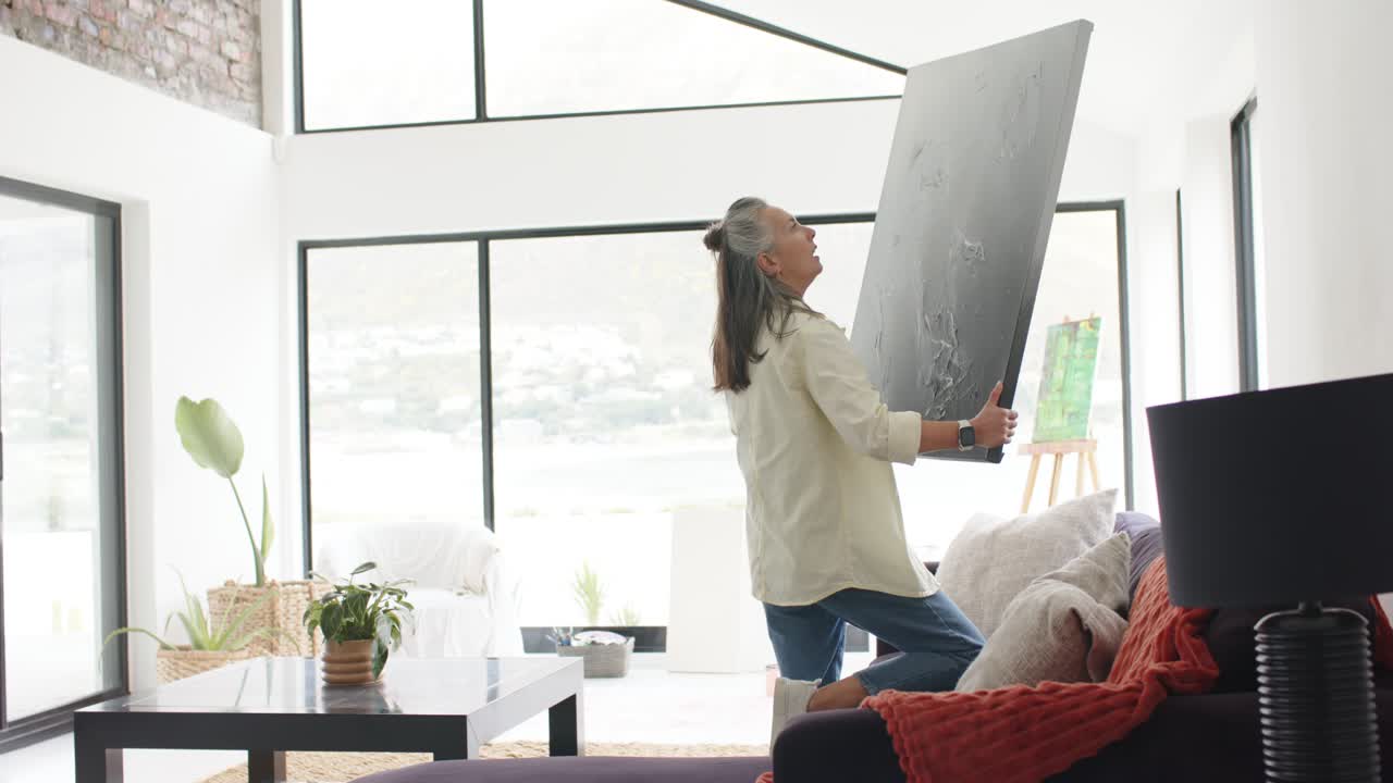 Moden hvid kvinde løfter et lærred hjemme, presser det mod væggen fra sofaen og hænger kunst op.