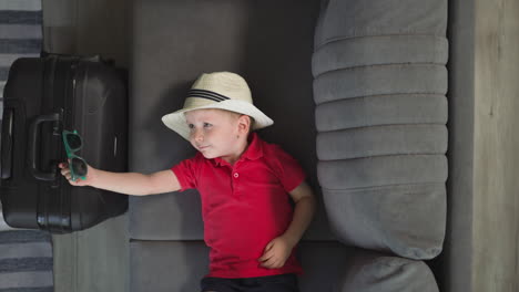 Little-traveler-in-hat-puts-sunglasses-on-big-black-suitcase