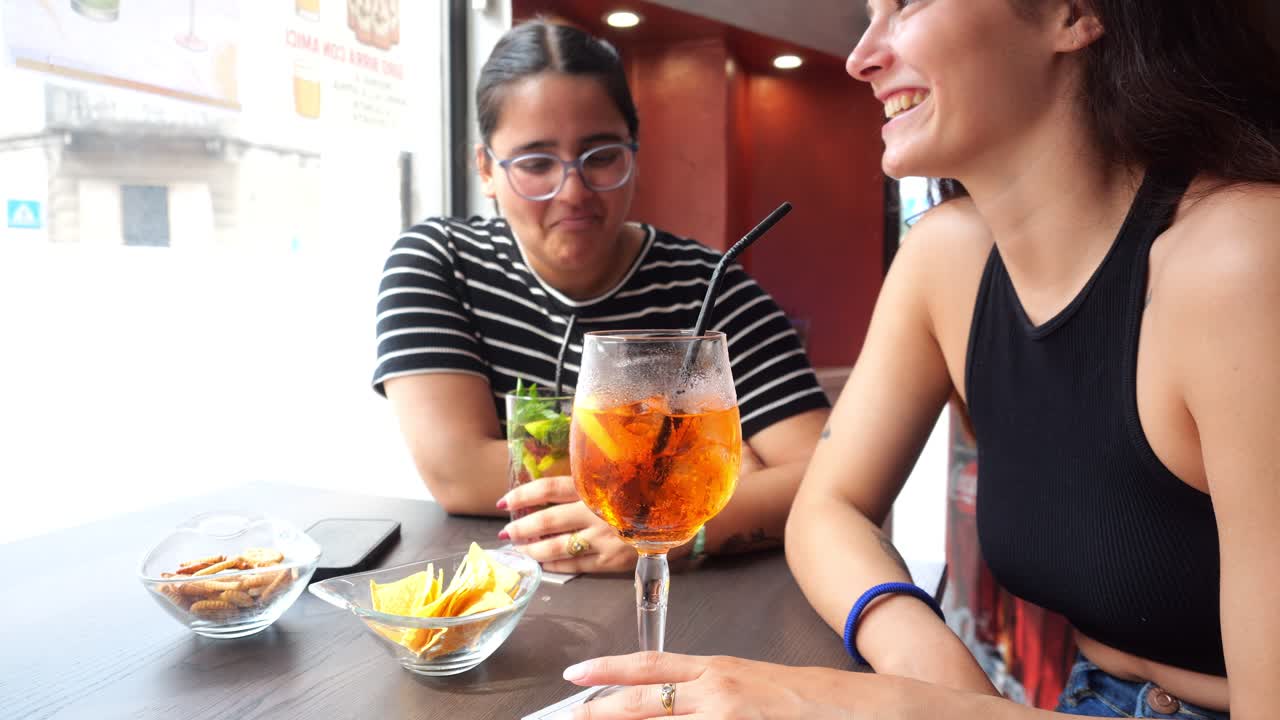Two young women sitting at a cafe table, enjoying drinks and snacks while browsing a smartphone, sharing laughs and moments of connection with each other in a cheerful atmosphere