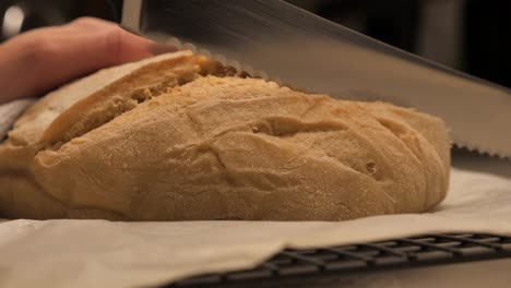 Persona-Cortando-Una-Hogaza-De-Pan-De-Masa-Agria-Recién-Horneada-Con-Un-Cuchillo-De-Cocina-Dentado-Afilado-En-La-Parte-Superior-De-Un-Banco