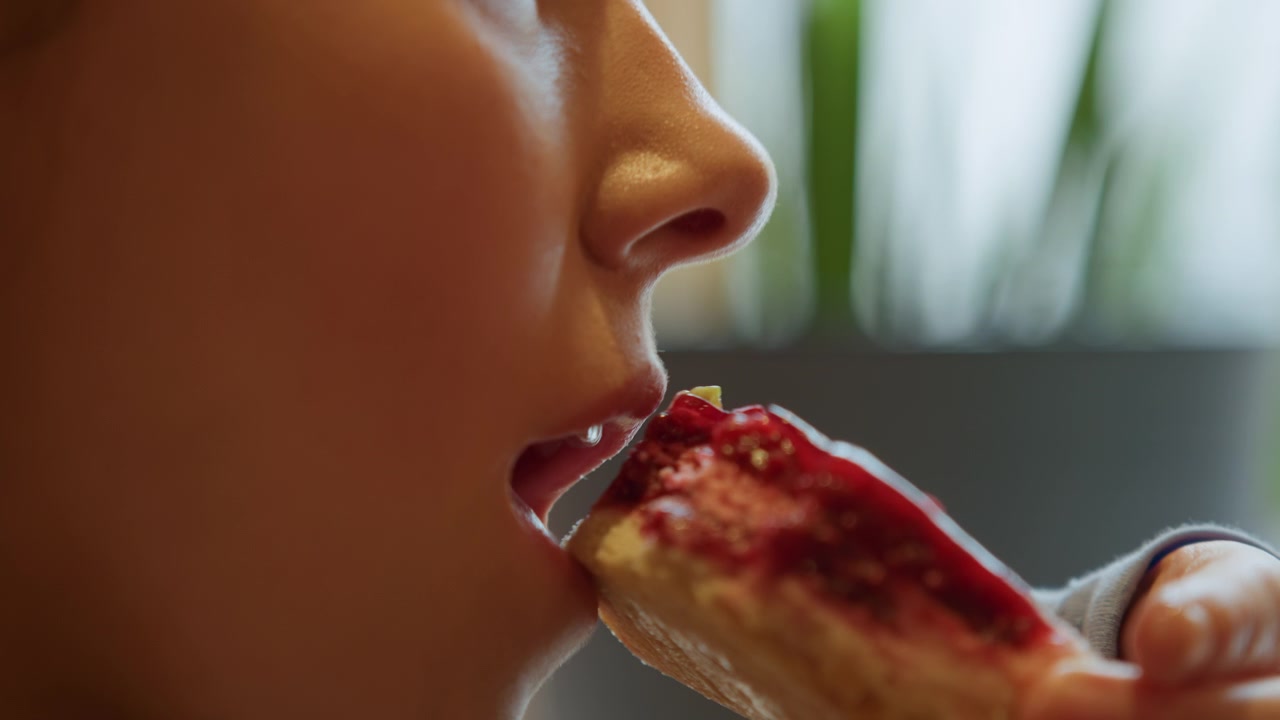 Close up of young woman eating food with visible texture detail. scene shows subject profile as woman takes bites focusing on her mouth and hand holding pastries or bread with red topping