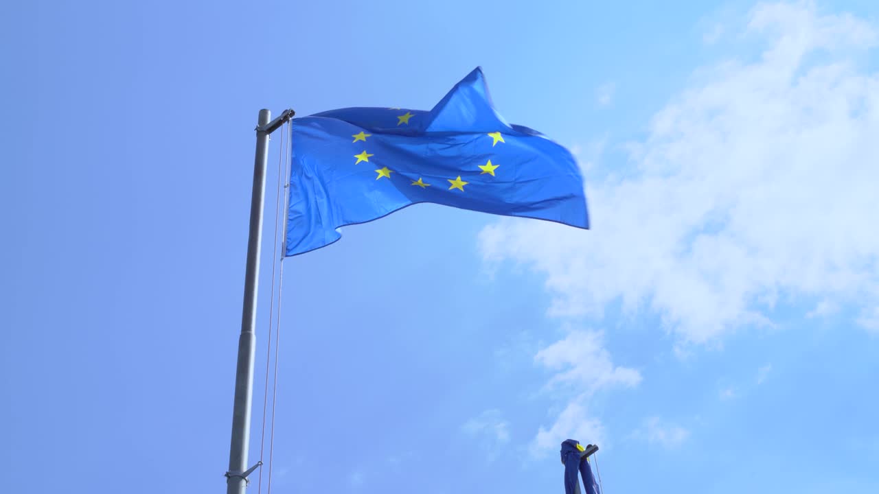 bandera de la unión política y económica europea con doce estrellas doradas en campo azul en un asta de metal ondea en el viento contra un cielo nublado en un día soleado