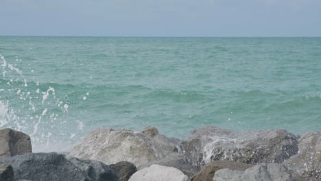 ocean waves crash against large coastal rocks, sending white spray into the air on a bright day