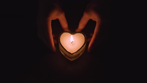 hands pushing romantic lit heart shaped white candle on black background into frame