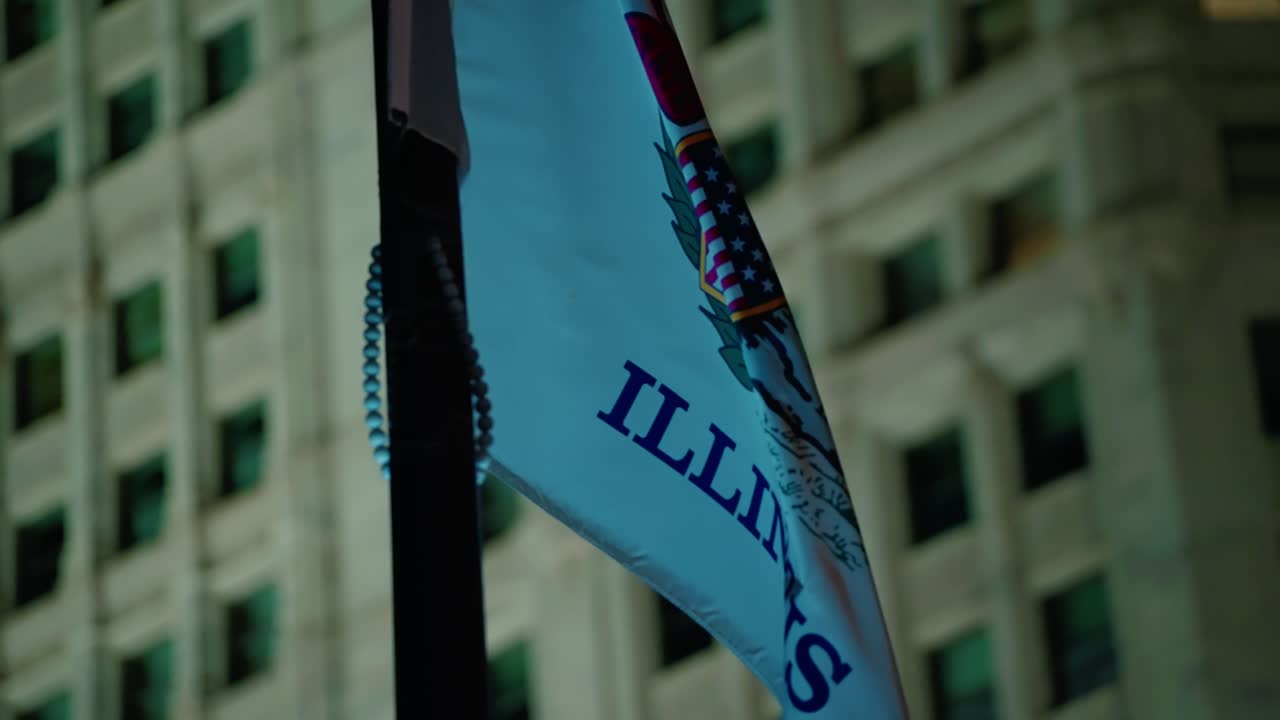 Primo piano della bandiera ufficiale dello stato dell'Illinois che sventola di notte contro lo sfondo di un oscuro edificio cittadino. Giorno delle elezioni a Chicago.