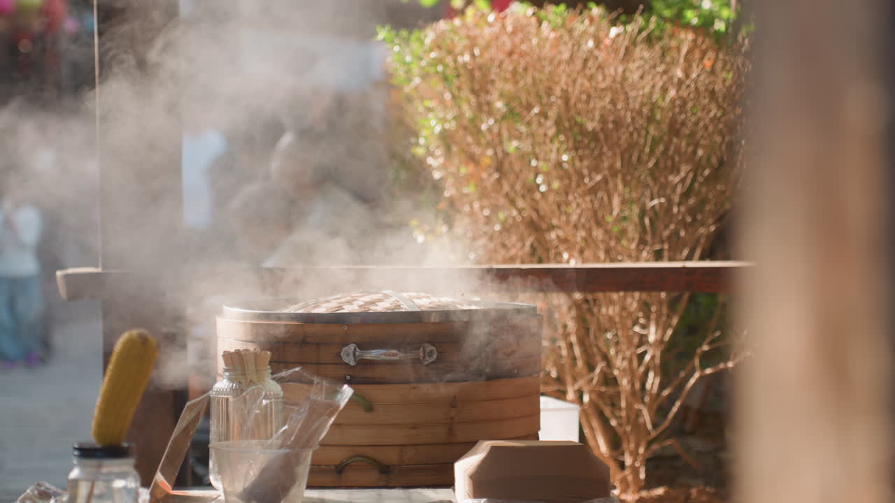 Dense steam billowing from bamboo steamer at busy market stall blurred crowd and festival energy in background, corn on stick and jars on counter, midday sunlight filtering through foliage, vendor