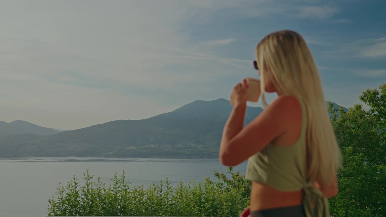 Blonde woman with long hair sips from a mug on a terrace while admiring panoramic views of Lake Maggiore and the surrounding mountains on a bright Italian summer day.