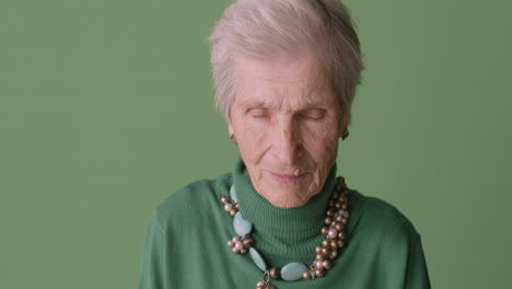 anciana rubia con el pelo corto usando suéter verde, aretes y collar posando sobre fondo verde mirando la cámara