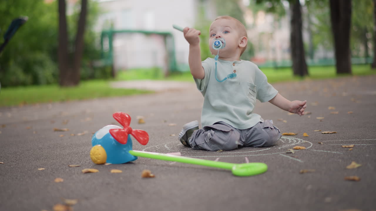 Bílé batole sedí na asfaltu, hraje si s větrníkem a lopatkou mezi spadaným listím, s dudlíkem v ústech. V pozadí jsou měkké stromy a kočárek, vytvářejíce poklidný a osamělý okamžik životního stylu.