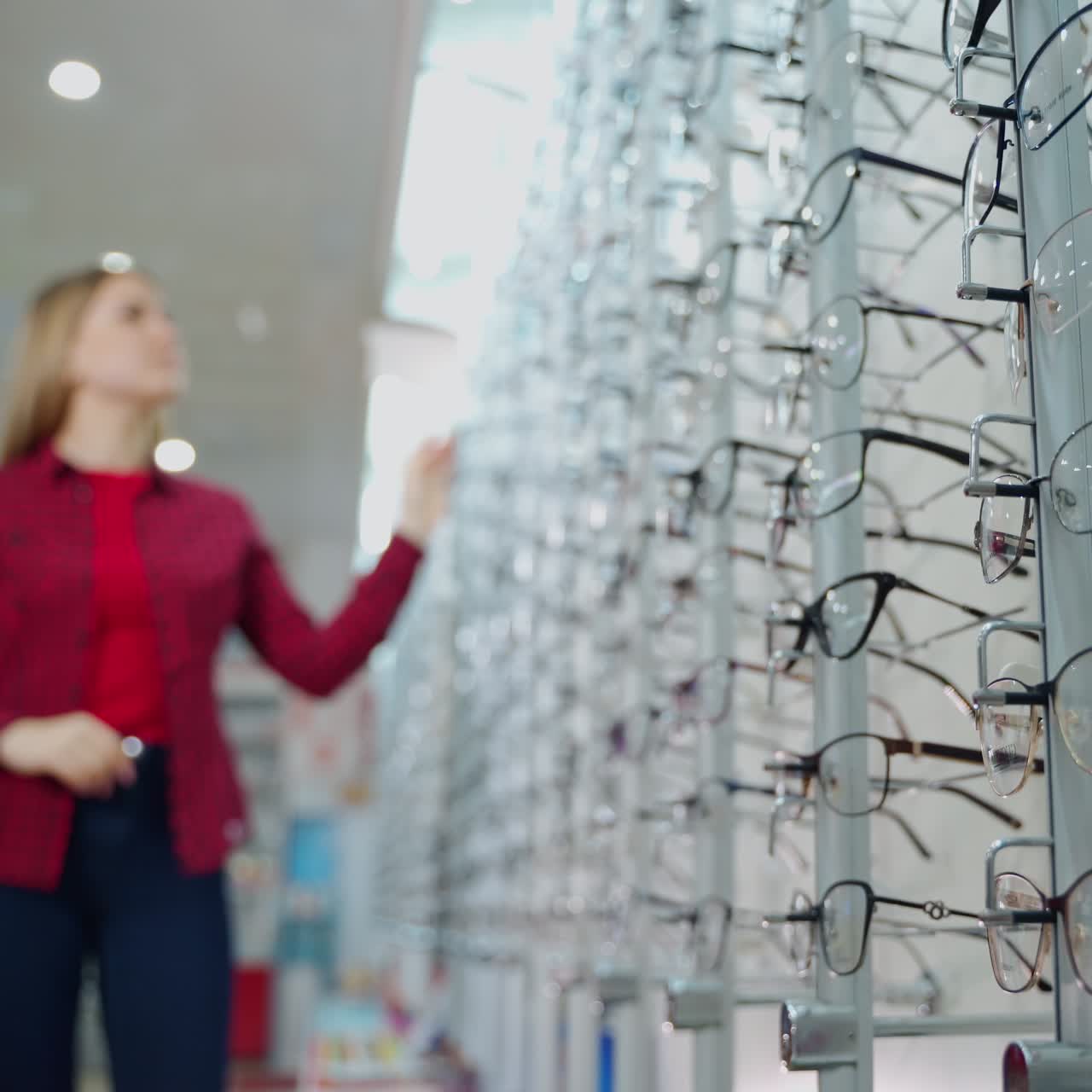 Fashionable eyeglasses in the optical store. Stand with new eyeglasses and a young woman choosing glasses. Vision concept.