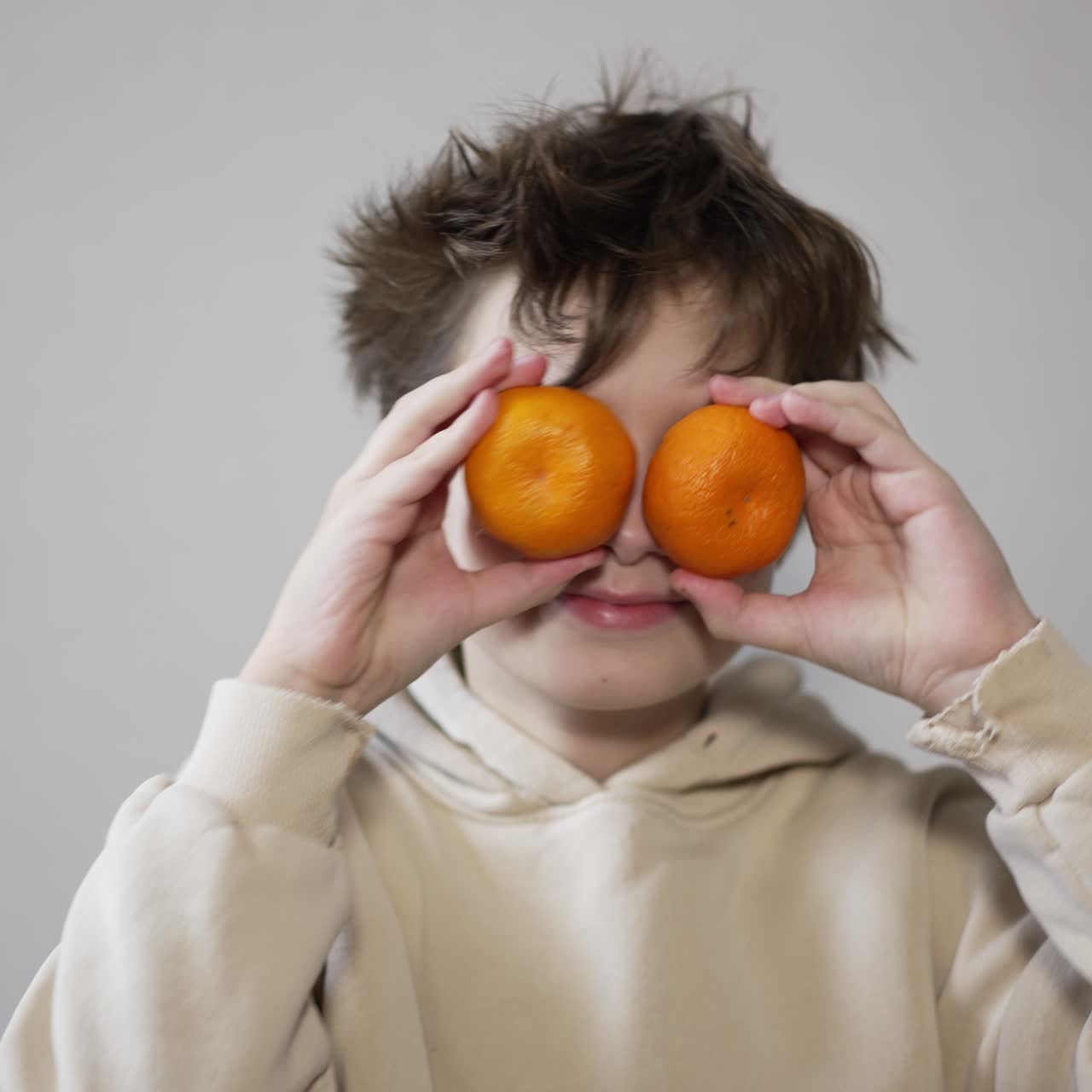 Cute brunet boy in hoodie puts two tangerines near his eyes. Caucasian teenager having fun. Close up