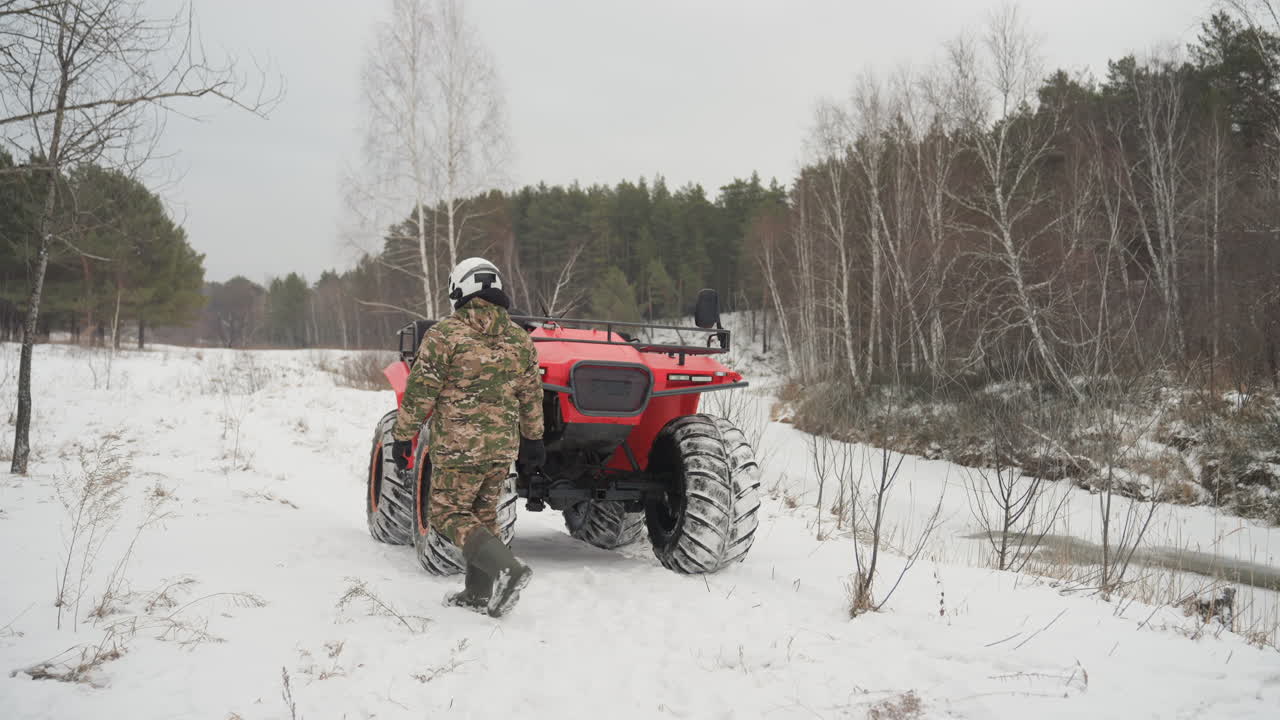 Un pilota mimetico si avvicina a un ATV rosso in una foresta di betulle innevata, ispeziona i pneumatici, sale sul veicolo e avvia il motore per un'avventura fuoristrada estrema, protetto dal casco. Cielo freddo e coperto, tracce visibili.