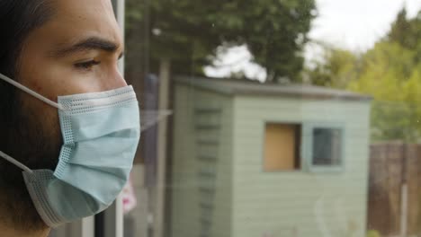 hombre mirando por una ventana con anhelo usando una máscara facial en cuarentena de cerca