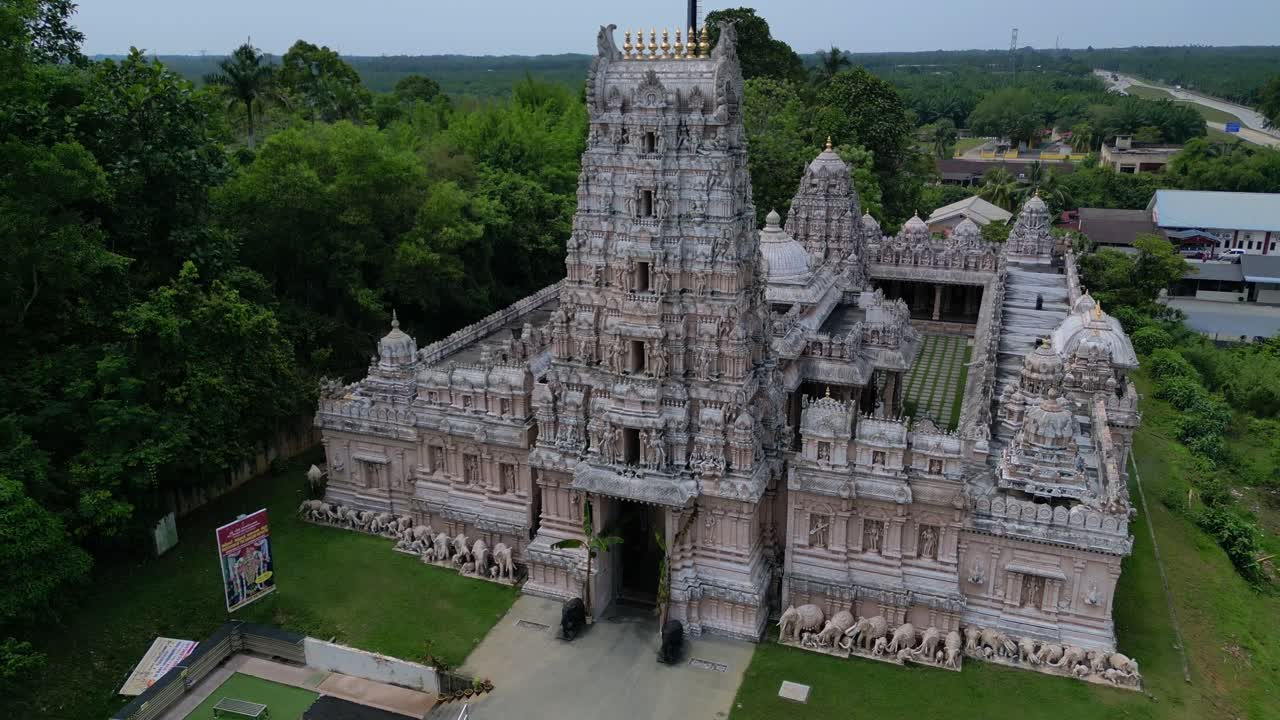 Ngoại thất đền Hindu trang trí công phu với tháp cổng gopuram cao chót vót, những chạm khắc tinh xảo và các bức tượng, bao quanh bởi cảnh quan xanh mát. Cảnh quay mượt mà từ trên không, drone bay lên tạo góc nhìn toàn cảnh.