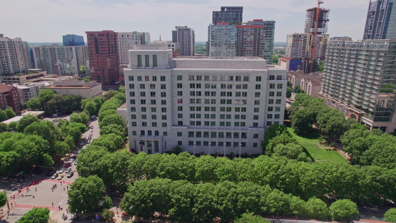 Vista aérea se aproximando do edifício de granito e mármore do Federal Reserve no centro de Atlanta, cercado por um exuberante dossel de árvores com o horizonte da cidade moderna ao fundo em um dia de verão ensolarado