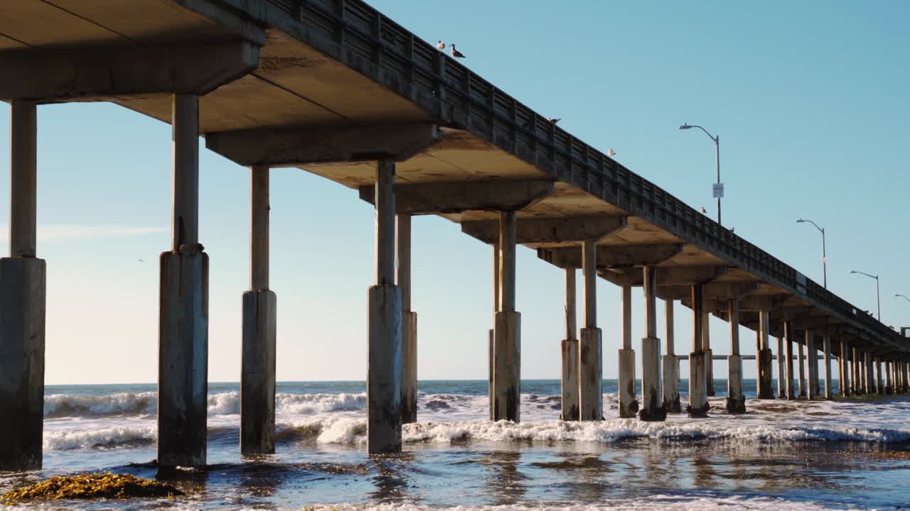 debajo de un paseo marítimo tropical como olas salpicando contra los pilares del muelle en un día sin nubes en el sur de california