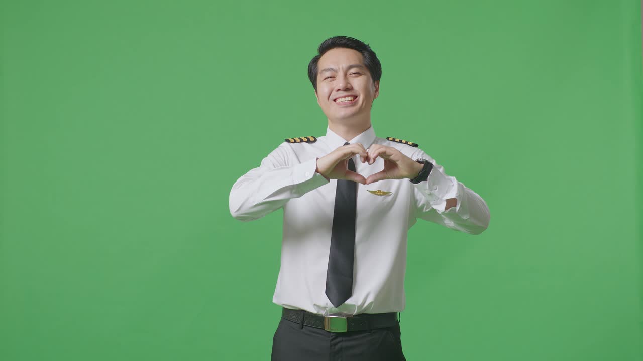 아시아 남성 조종사가 녹색 스크린 배경에서 심장 몸짓을 하고 미소를 짓고 있습니다.